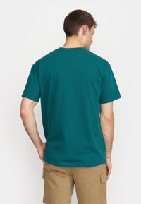 Camiseta de manga corta en color azul verdoso, hecha de una tela suave, con un cuello redondo clásico y un corte relajado, con detalles de costura visibles, combinada con pantalones cortos color tostado.