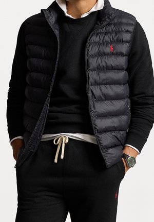 Man wearing black quilted vest over black sweatshirt and black sweatpants with white drawstring, hands in pockets, and wristwatch on left wrist.