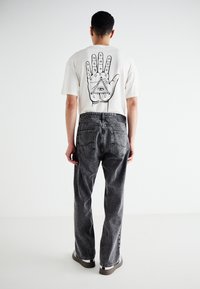 White cotton t-shirt with black graphic of a hand and eye on the back, paired with high-waisted gray denim jeans and black shoes.