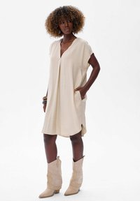 Beige, jurk tot de knie met korte mouwen, V-hals, zijzakken en een vloeiende pasvorm. Gecombineerd met lichtbeige cowboylaarzen.