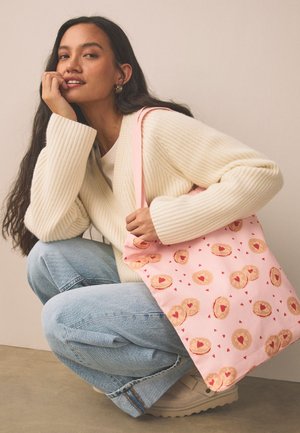 Kobieta z długimi ciemnymi włosami, ubrana w kremowy sweter i jasnoniebieskie dżinsy, kuca i trzyma różową torbę z wzorami serc i ciasteczek.