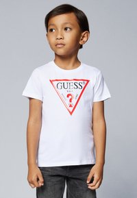 Weißes Baumwoll-T-Shirt mit einem roten und schwarzen dreieckigen Logo und dem Text "GUESS U.S.A.". Kurze Ärmel und ein Rundhalsausschnitt. Graue Jeans.