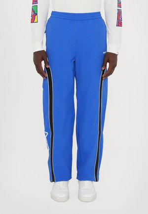 Blue track pants with black side stripes and white accents, featuring an elastic waistband and a loose fit. Paired with white sneakers.