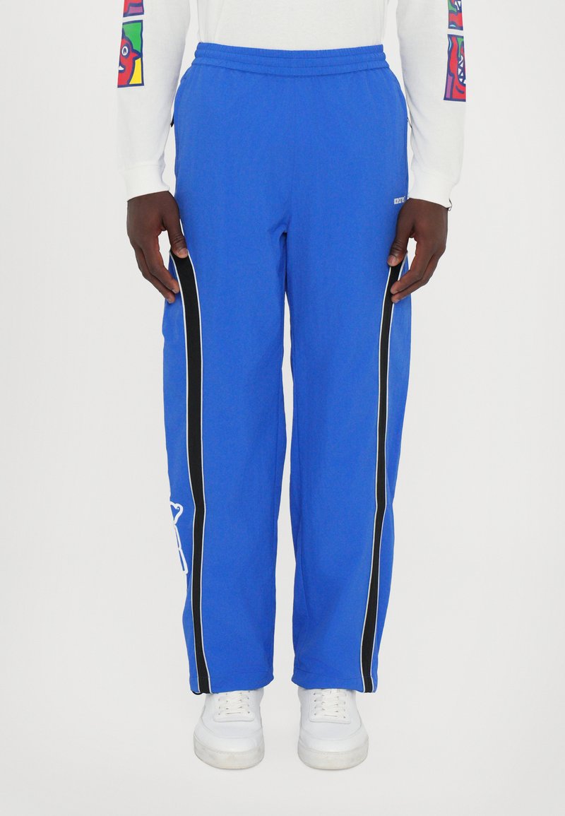 Blå joggebukser med svarte sideflater og hvite detaljer, med elastisk midjebånd og løs passform. Kombinert med hvite joggesko.