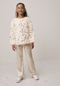 Cream fuzzy sweater with shimmering embellishments, paired with sparkly gold wide-leg pants and white sneakers. Simple background.