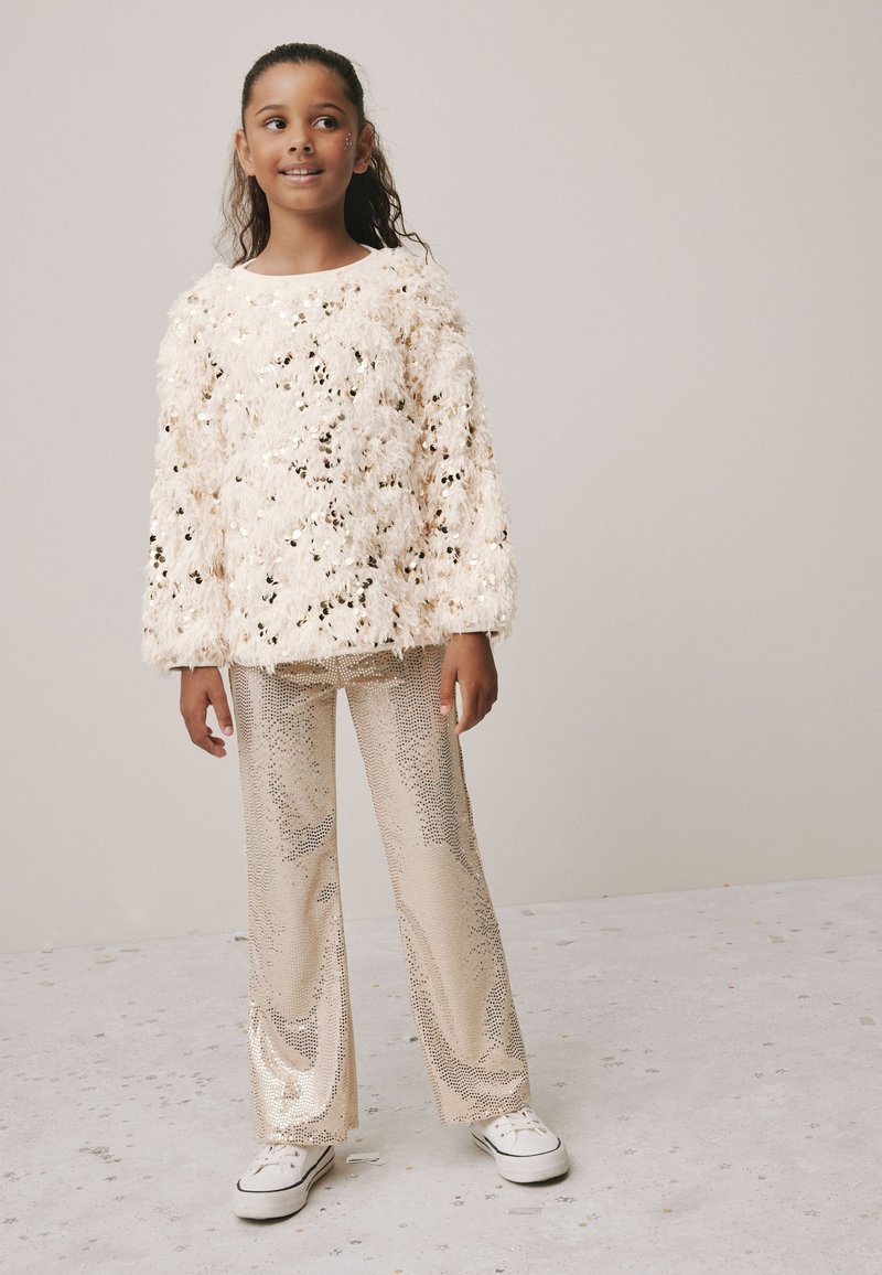Cream fuzzy sweater with shimmering embellishments, paired with sparkly gold wide-leg pants and white sneakers. Simple background.