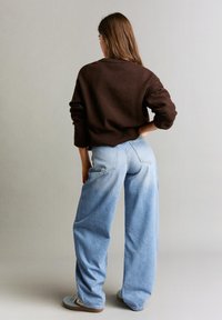 Donna in piedi di spalle alla fotocamera, che indossa un maglione marrone scuro, jeans a gamba larga azzurri e sneakers azzurre.