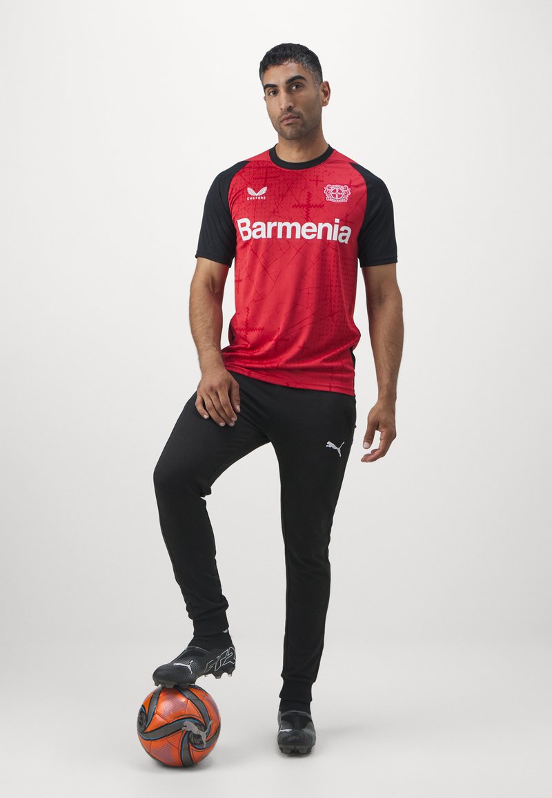 Camisa desportiva vermelha e preta com o logo "Barmenia", acompanhada por calças pretas. Usando sapatilhas de futebol pretas com uma bola de futebol laranja aos pés.