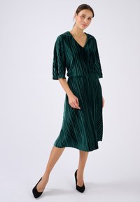 Grünes Samtkleid mit vertikalen Streifen, V-Ausschnitt und drei Viertelärmeln; kombiniert mit schwarzen Absätzen. Sanfte Textur und fließende Silhouette.