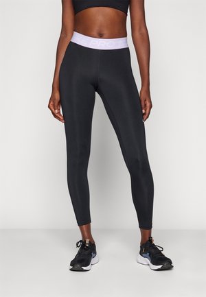 Modelka w czarnych legginsach Nike Pro z wysokim stanem i jasnofioletowym paskiem oraz czarnych butach sportowych, stojąca na jednolitym tle.