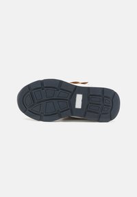 Suela de zapato que presenta un diseño de goma negra gruesa con una superficie texturizada y patrones de banda de rodadura definidos para tracción. Se pueden ver acentos marrones.