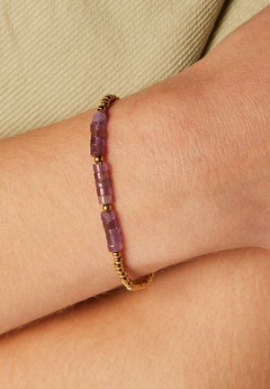 Gold bracelet featuring small purple rectangular beads and round gold accents, highlighting a textured, beaded design on a wrist.