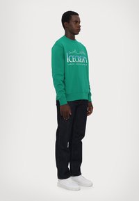 Grøn sweatshirt med hvid "ICECREAM" grafik, parret med mørk denim jeans og hvide sneakers. Sweateren har ribbede manchetter og kant.