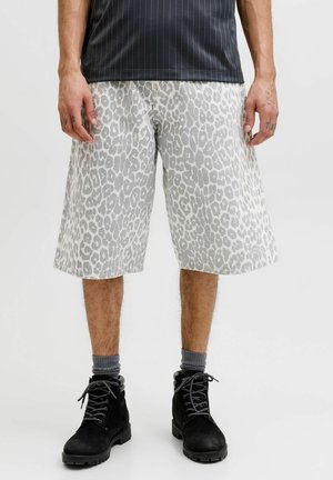 Mężczyzna ubrany w szare i białe szorty o długości do kolan w panterkę, czarne buty sznurowane oraz ciemne skarpety, stojący na jednolitym tle.