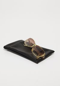 Solfanger med en gjennomsiktig gul ramme og gradient brune glass, plassert på en svart lærpung med «QUAY» preget.