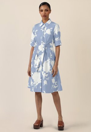 Frau trägt ein knielanges, hellblaues Kleid mit weißen Blumenmustern, kurzen Ärmeln, vorderem Knotendetail und braunen Plateau-Sandalen.
