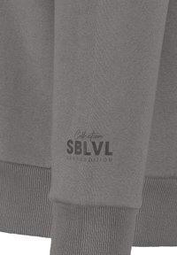 Grauer Sweatshirt mit gerippten Bündchen, glatter Textur und bedrucktem Logo "SBLVL URBAN EDITION" nahe dem Saum.