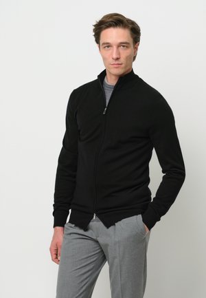 Maglione nero con zip e colletto alto, design aderente, abbinato a pantaloni grigi. Texture liscia, dettagli costruttivi sottili e un look classico.