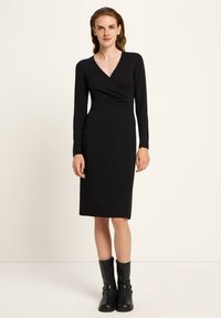 Eine Frau mit langen Haaren, die ein schwarzes langärmeliges Wickelkleid und schwarze Stiefel bis zur Mitte der Wade trägt, steht vor einem schlichten hellen Hintergrund.