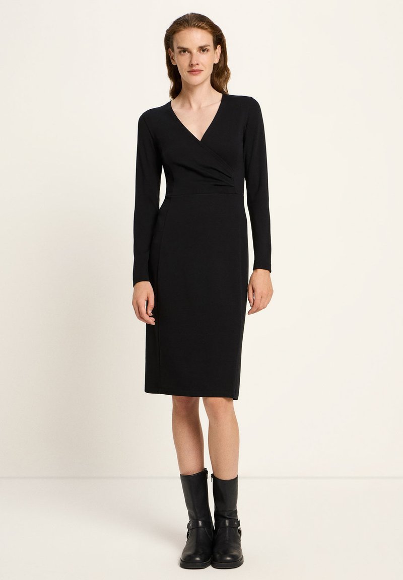 Eine Frau mit langen Haaren, die ein schwarzes langärmeliges Wickelkleid und schwarze Stiefel bis zur Mitte der Wade trägt, steht vor einem schlichten hellen Hintergrund.