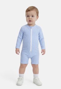 Malček stoji, oblečen v modro-belo romper z navpičnimi črtami in dolgimi rokavi, z zadrgo spredaj, belimi nogavicami in belimi supergami.