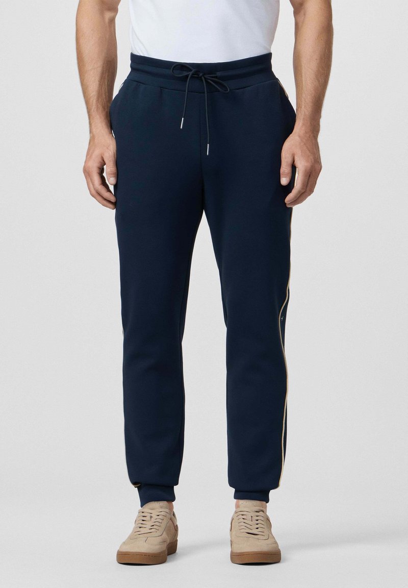 Uomo che indossa pantaloni jogger blu navy con strisce laterali, maglietta bianca e sneakers beige, in piedi davanti a uno sfondo semplice.