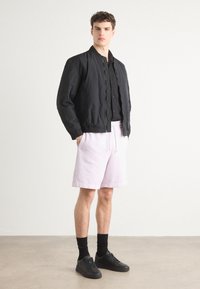 Sort bomberjakke med ribbet krave, båret over en sort skjorte med knapper. Lyserøde shorts og sorte sneakers fuldender looket.