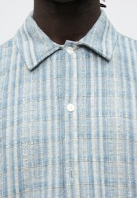 Inimese lähivõte, kes kannab helesinist ja valget triibulist kootud särk, millel on krae ja valged nupud, mis on ülaosas kinni kinnitatud.