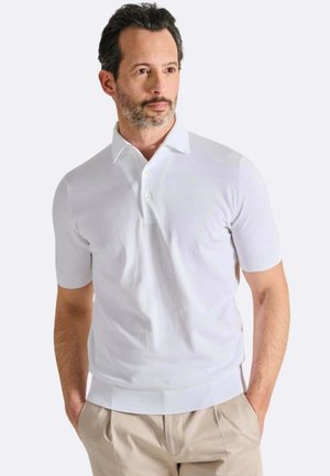 Uomo con capelli corti scuri e barba che indossa una polo bianca a maniche corte e pantaloni beige, con le mani nelle tasche, che guarda di lato.