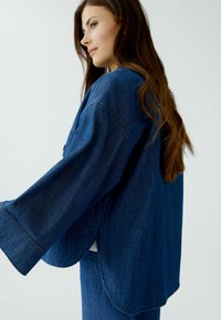 Donkerblauwe denimjas in een oversized pasvorm met brede mouwen, een kromme zoom en zichtbare stikseldetails. Zachte textuur, casual ontwerp.
