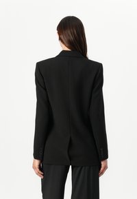 Musta räätälöity takki terävällä kauluksella, rakenteellisilla harteilla ja napitettavilla hihoilla, jossa on yksittäinen tuuletusaukko selkäpuolella liikkumisen helpottamiseksi.