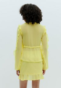 Geel doorschijnende jurk met ruches op de mouwen en de zoom, gelaagd ontwerp met een aansluitende taille, en een zachte, lichte textuur.