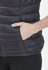 Chaleco de plumas negro con diseño acolchado horizontal, bolsillo con cremallera y exterior suave y brillante. Mano situada en el bolsillo.