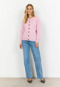 Cardigan rosa con scollo rotondo, bottoni di legno e maniche lunghe, abbinato a jeans azzurri chiari e tacchi a strisce nere e bianche.