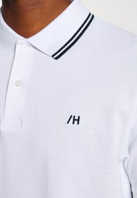 Λευκό πόλο μπλούζα με μαύρες ρίγες στο γιακά, με ένα μικρό μαύρο κεντητό λογότυπο "/H" στο στήθος. Υφασμάτινη υφή.