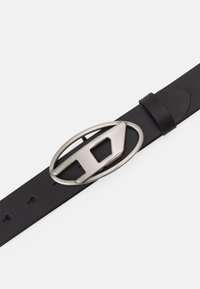 Ceinture en cuir noir avec une boucle ovale en argent arborant une découpe géométrique. Texture lisse, avec des trous d'ajustement le long de la sangle.