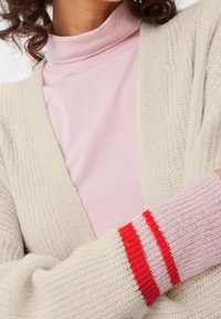 Krémový pletený kardigan s širokými rukávy, zdobený růžovou ribbed manžetou s červenými a bílými pruhy, vrstvený přes světle růžový rolák.