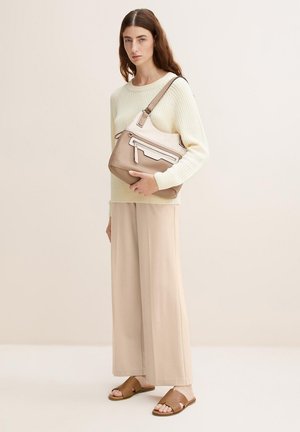 Borsa a spalla in pelle tan con finitura liscia, dettaglio della cerniera bianca e tracolla regolabile. Il modello indossa un maglione crema e pantaloni beige.
