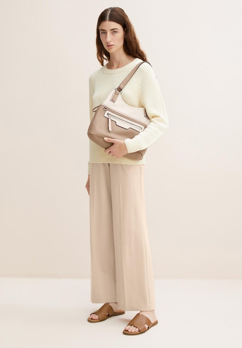 Bolso de hombro de cuero marrón claro con un acabado suave, detalle de cremallera blanca y correa ajustable. El modelo lleva un suéter crema y pantalones beiges.