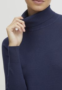 Mörkblå tubhalsad tröja i mjukt stickat material med ribbade detaljer vid kragen och ärmarna. Närbild av en hand som lätt rör vid kragen.