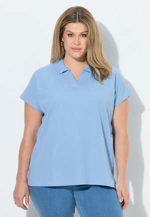 Kobieta z blond włosami do ramion, ubrana w jasnoniebieską koszulkę polo z krótkim rękawem i niebieskie dżinsy, stoi przy białej ścianie.