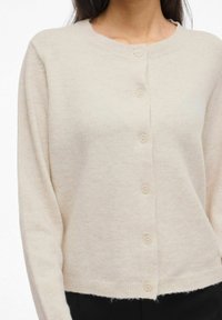 Bēšs adīts kardigans ar apaļu kakla izgriezumu, garām piedurknēm un sešām pogām priekšpusē, nedaudz īsāka apakšmala.