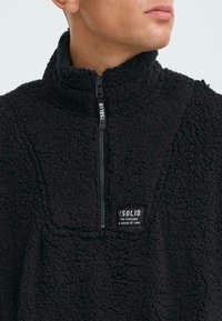 Schwarzer, mit Reißverschluss versehener Fleece-Pullover mit hohem Kragen, strukturierter Oberfläche und verstärkten Nähten; verfügt über ein kleines Markenpatch auf der Vorderseite.