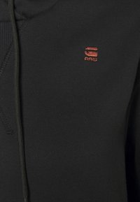 Černá mikina s texturovanou tkaninou, která má na hrudi malé červené vyšívané logo a nastavitelnou kapuci se šňůrkou.