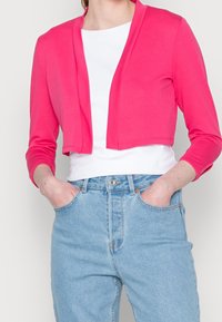 Pink cropped cardigan with long sleeves, soft fabric, large open front, paired with a white top and light blue high-waisted jeans.