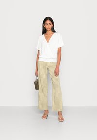 Weißes Wickeloberteil mit kurzen Ärmeln, kombiniert mit weit geschnittenen Hosen in grün-gelbem Karomuster. Modell hält eine kleine olivgrüne Tasche.