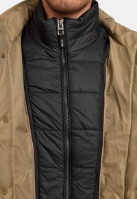 Schwarze Pufferjacke mit gestepptem Muster, ausgestattet mit hohem Kragen und durchgehendem Reißverschluss, unter einem tanfarbenen Außenmantel mit Knöpfen getragen.