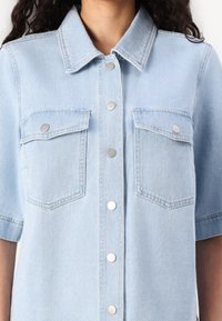 Camisa de ganga azul claro com colarinho, mangas curtas e dois bolsos no peito, apresentando botões de pressão prateados na parte da frente e detalhes de costura sutis.