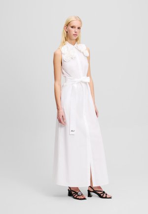 Witte, mouwloze midi-jurk met bloemapplicaties op de kraag, strikceintuur, knoopsluiting aan de voorkant en een zijsplit. Zwarte, smalle hakken.