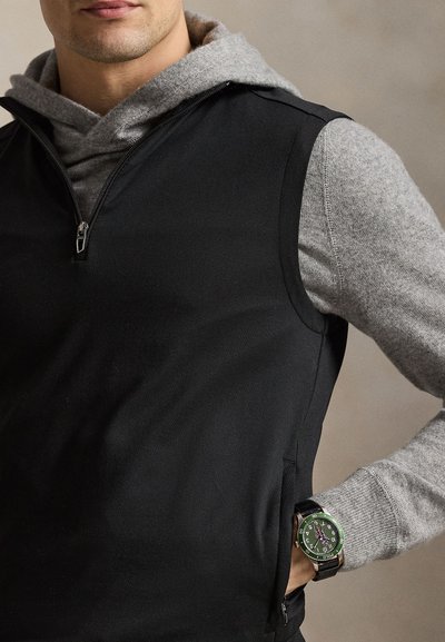 Άνδρας που φορά γκρι φούτερ με κουκούλα κάτω από μαύρο γιλέκο χωρίς μανίκια, με ρολόι με πράσινο καντράν στον αριστερό του καρπό.